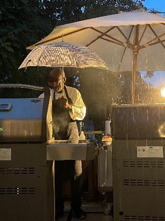 Das Foto zeigt einen Koch, der unter einem Regenschirm am Grill steht.